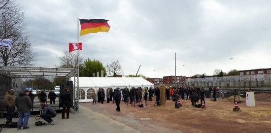 Die Gäste füllen langsam den Platz für den feierlichen Baubeginn des Stellinger Deckels. Rechts auf dem Platz der vorbereitete Startknopf für den Baubeginn, hinten auf der anderen Autobahnseite vor den Häusern ein schon fertiges Teilstück der provisorischen, oben durchscheinenden, Lärmschutzwand.