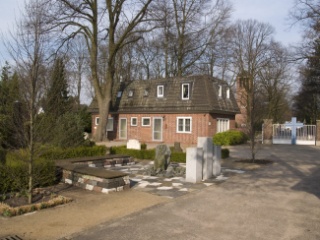 Blick von der Kapelle auf den Vorplatz mit den Bänken, dahinter das Gebäude der ehemaligen Friedhofsverwaltung.